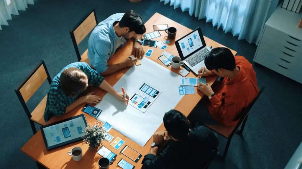 Equipe de trabalho reunida em uma mesa e analisando documentos e gráficos. Também há um notebook aberto em cima da mesa. Há na imagem três homens e uma mulher Todos são brancos.