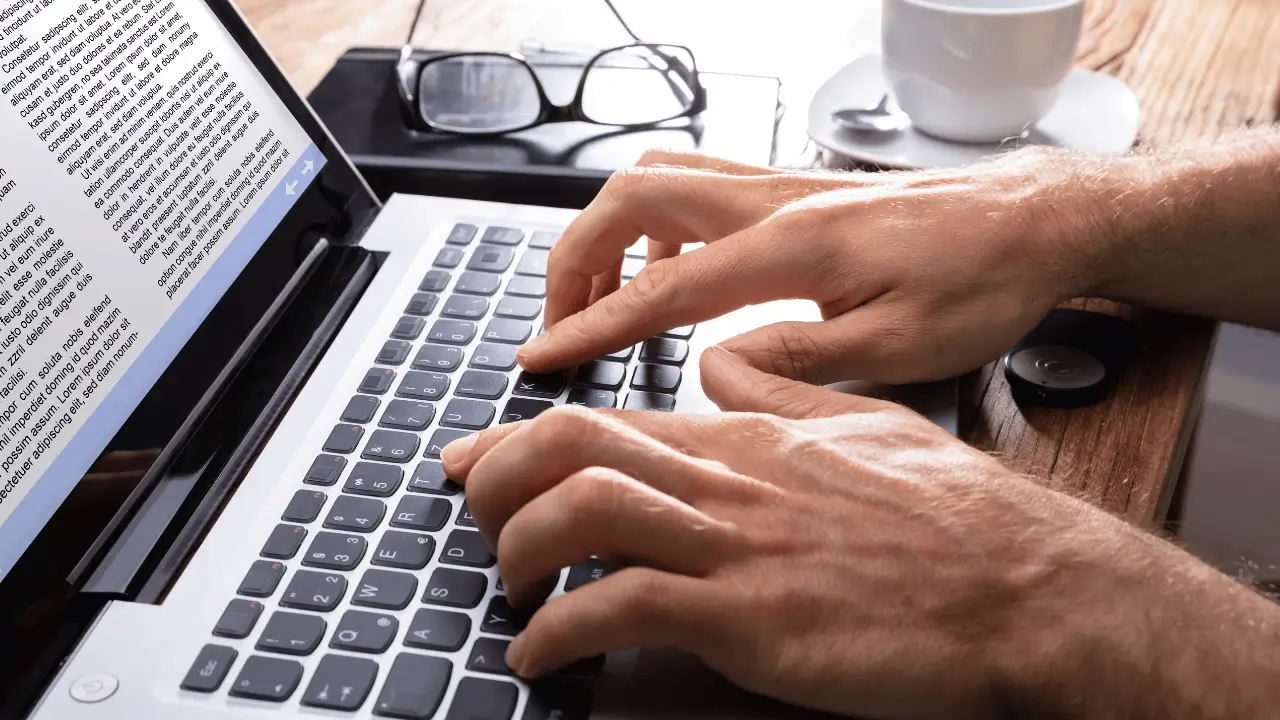 Homem digitando em um notebook, que na tela mostra um bloco de texto. Aparece apenas as mãos do homem. Ele é branco.