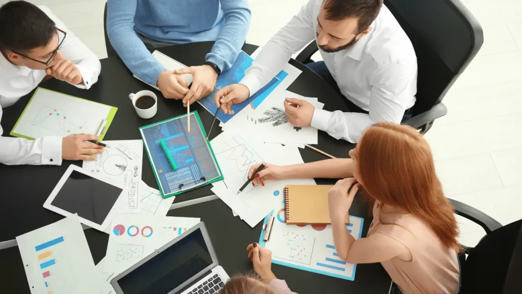 Equipe reunida em uma mesa, com muitas folhas e um notebook em cima da mesa. Há dois homens e duas mulheres. Todos são brancos e vestem roupas sociais