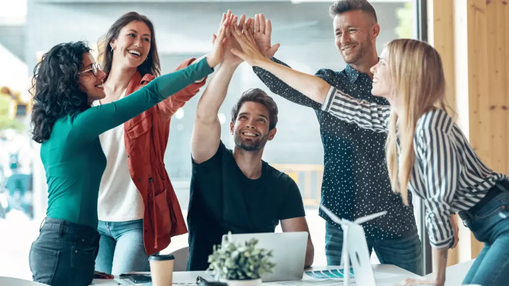 Equipe de trabalho unindo as mãos, com os braços esticados para cima. São cinco pessoas, sendo dois homens e duas mulheres. Todos são brancos.