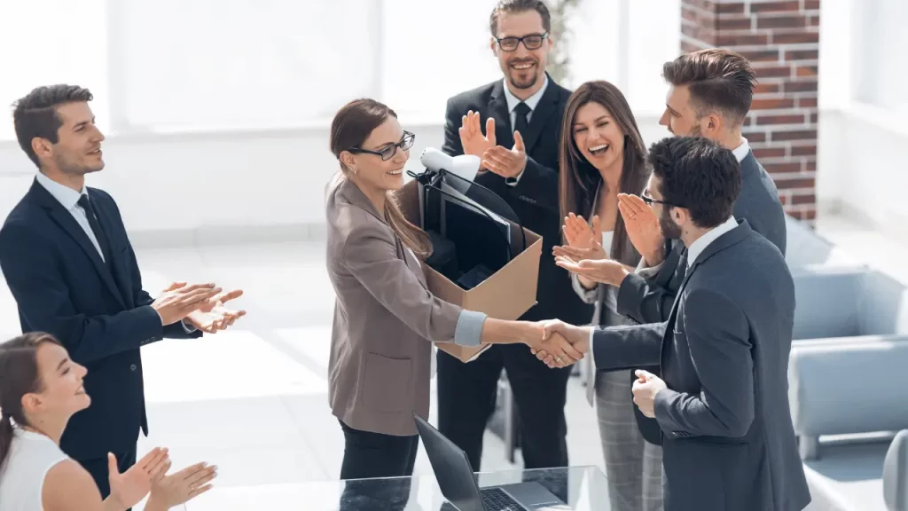 Equipe de trabalho reunida, interagindo  de maneira descontraída. No centro da imagem, há uma mulher com uma caixa de papelão em baixo d eu dos braços, cumprimentando um homem. 