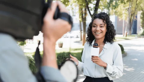 Imagem mostra uma repórter de TV, com o microfone na mão e um câmera filmando ela. A mulher é negra e veste camisa social branca.