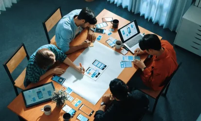 Equipe de trabalho reunida em uma mesa e analisando documentos e gráficos. Também há um notebook aberto em cima da mesa. Há na imagem três homens e uma mulher Todos são brancos.