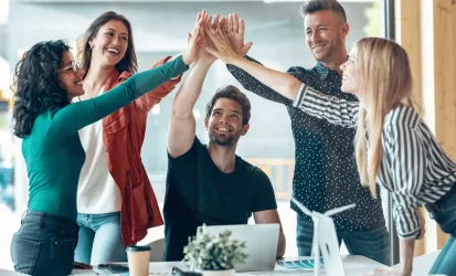 Equipe de trabalho unindo as mãos, com os braços esticados para cima. São cinco pessoas, sendo dois homens e duas mulheres. Todos são brancos.